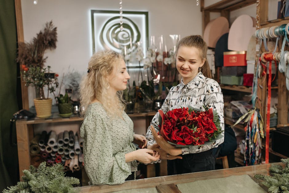 A young woman with long, light brown hair tied back, smiling as she receives a vibrant bouquet of red roses, lilies, and other mixed flowers wrapped in green foliage and light brown paper from a femal
