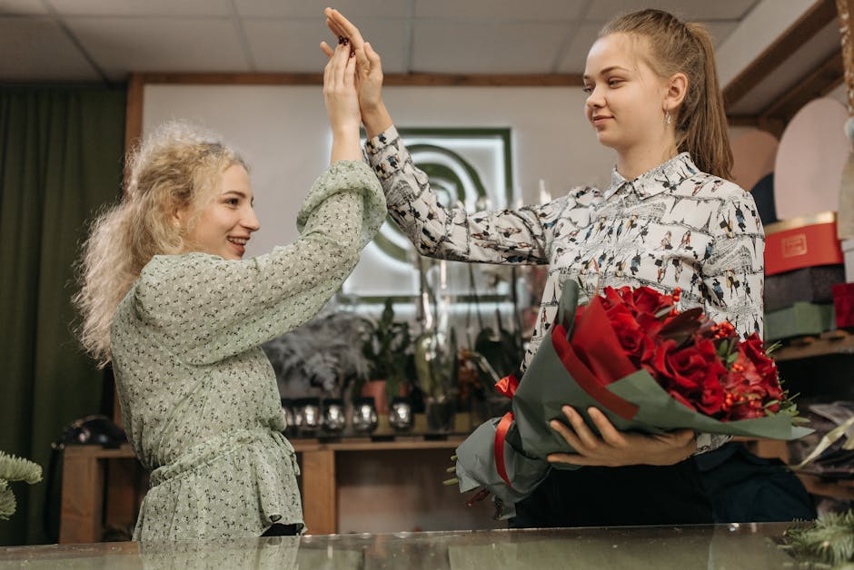 A florist and a customer in a flower shop exchanging a high-five, with the customer holding a bouquet of vibrant red roses wrapped in dark green paper. The florist, a young woman with long hair tied b