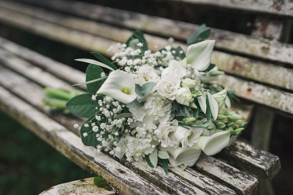 A bouquet of fresh white flowers arranged elegantly on an aged wooden park bench outdoors. The floral arrangement includes white calla lilies, roses, and spray roses, accented with delicate sprigs of 