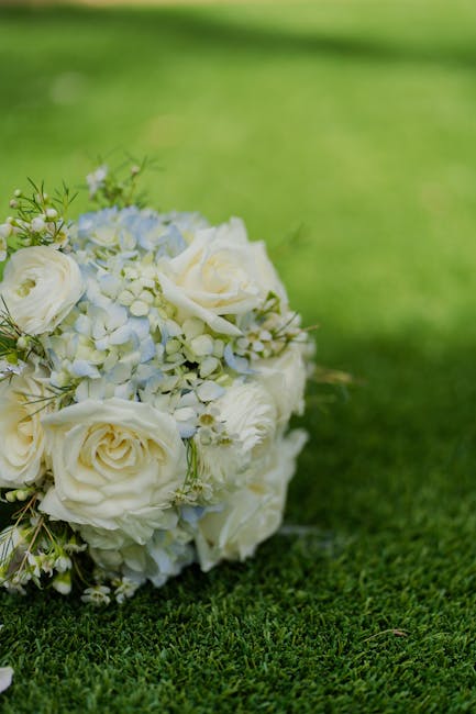 A close-up view of a bridal bouquet arranged with creamy white roses, delicate white hydrangeas, and sprigs of greenery, resting on lush green grass. The bouquet is wrapped in elegant white tissue pap