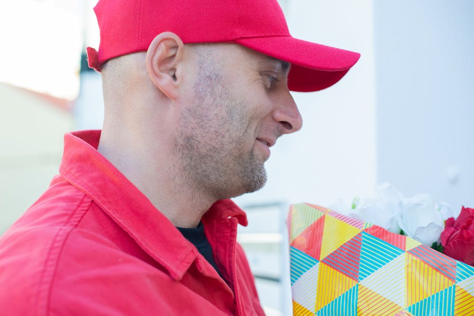 A man wearing a red cap and a red jacket is holding a colorful floral gift bag with a geometric pattern in shades of yellow, blue, red, and green, filled with white and pink flowers. He is outdoors in